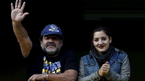 Maradona mit seiner Tochter Diego Maradona mit erhobener Hand und eine junge Frau stehen zusammen vor einem dunklen Hintergrund.