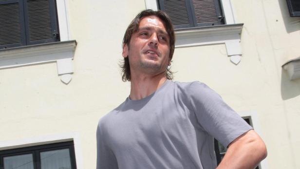 Ein Mann mit langen Haaren und einem grauen T-Shirt steht vor einem Gebäude.
