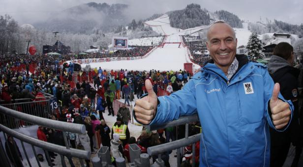 Ein Mann in einer blauen Jacke gibt vor einem Skirennen mit vielen Zuschauern die Daumen hoch.