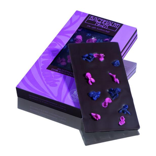 Eine Tafel dunkler Schokolade von Bachhalm mit rosa und blauen Dekorationen liegt neben der Verpackung.