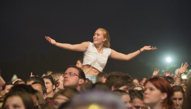 Eine Frau sitzt auf den Schultern einer Person bei einem Konzert und streckt die Arme aus.