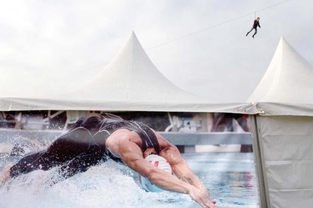Ein Werbeplakat eines Schwimmers vor einem weißen Zelt, während eine Person an einem Seil über sie hinwegfliegt.