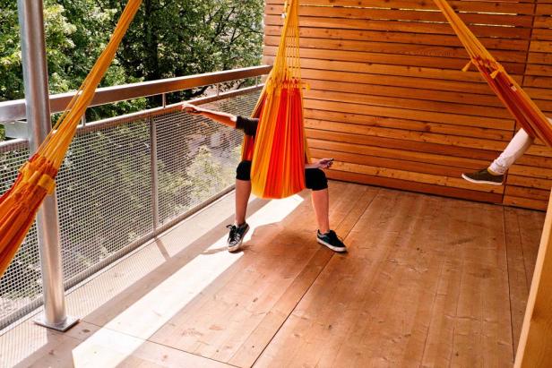 Eine Person sitzt in einer orangefarbenen Hängematte auf einem Holzbalkon.