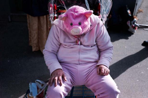 Eine Person mit rosa Trainingsanzug und Schweinsmaske sitzt auf einem Stuhl.