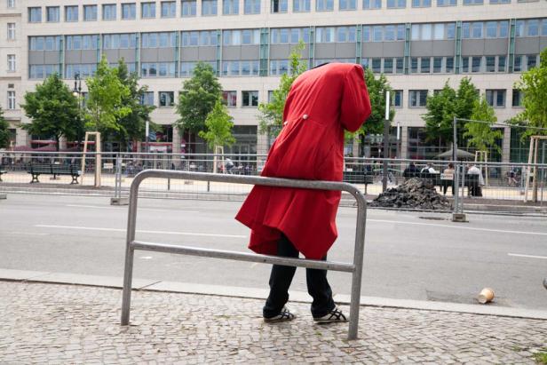 Eine Person in einem roten Mantel lehnt an einer Absperrung auf einer Straße.