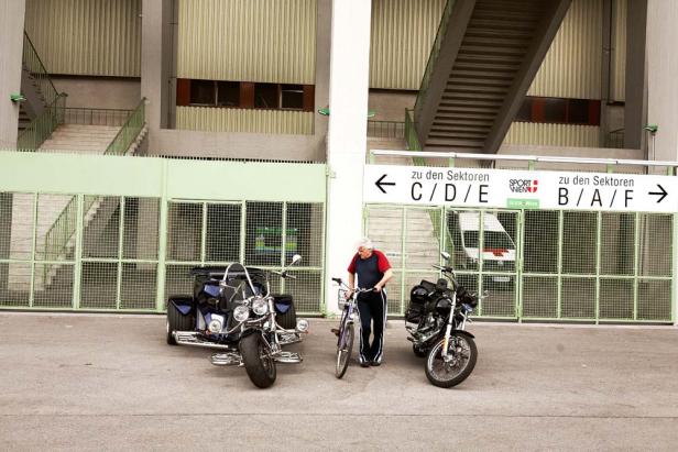Ein Mann mit Fahrrad steht vor dem Eingang zu einem Stadion, flankiert von einem Trike und einem Motorrad.