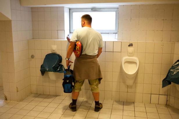Ein Mann steht mit Helm und Tasche in einem gefliesten Urinalraum.
