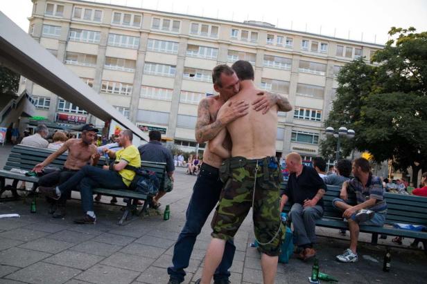 Zwei Männer umarmen sich auf einem öffentlichen Platz, während andere auf Bänken sitzen.