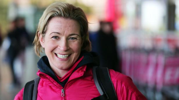 Eine lächelnde Frau mit Rucksack und pinkfarbener Jacke steht im Vordergrund.