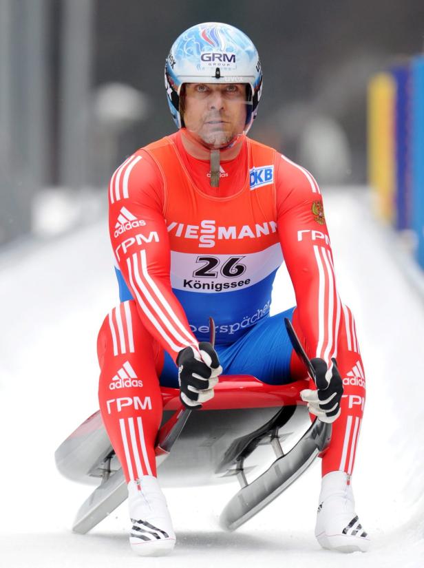 Ein Rennrodler in roter und blauer Rennkleidung sitzt auf seinem Schlitten.