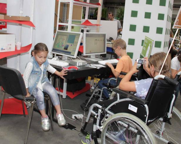 Drei Kinder sitzen an einem Tisch und benutzen Computer, eines davon im Rollstuhl.