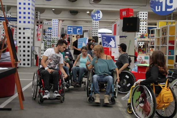 Eine Gruppe von Menschen sitzt in Rollstühlen in einer Halle mit verschiedenen Ständen.