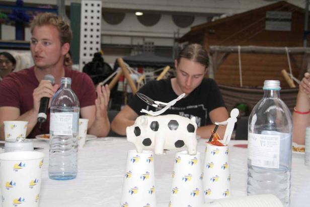 Ein Mann spricht in ein Mikrofon, während ein anderer eine Dekoration auf einem Tisch betrachtet.