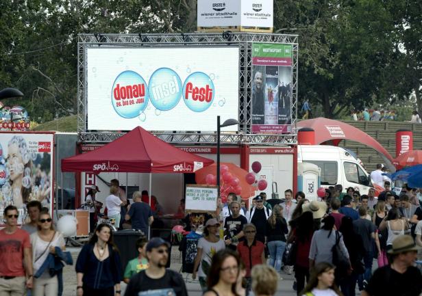 Menschen besuchen das Donauinselfest vor einer Bühne mit der Aufschrift „Donauinselfest 26. bis 28. Juni“.