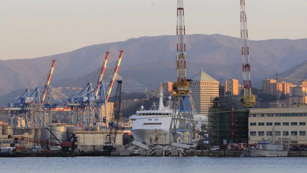 Ein Schiff liegt im Hafen von Genua, umgeben von Kränen und Gebäuden.