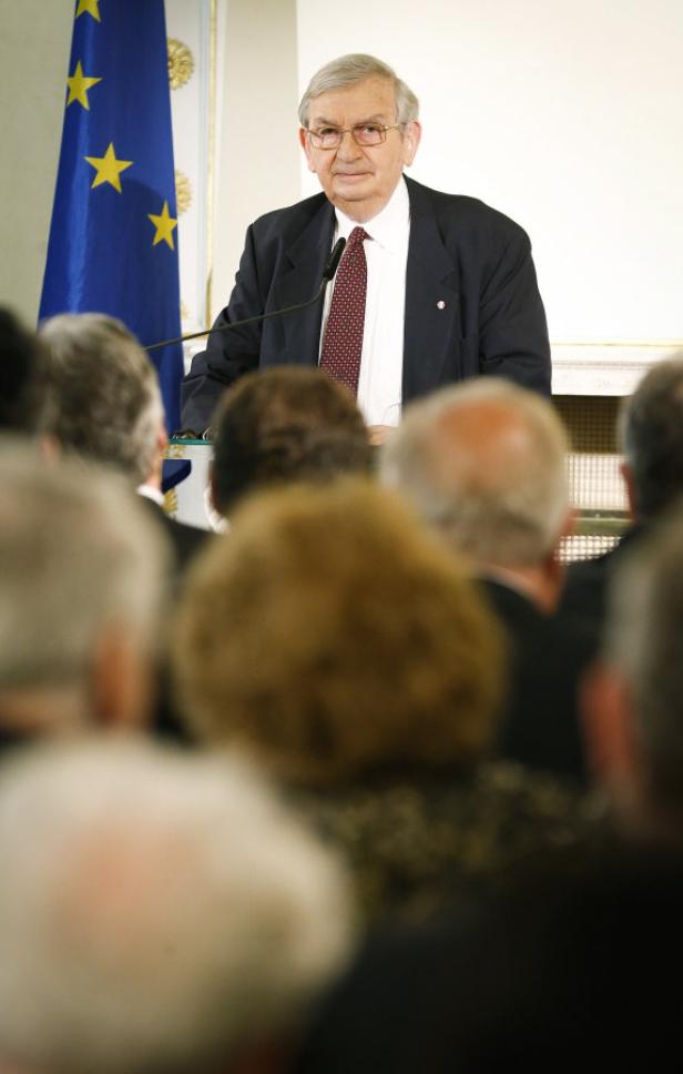Ein älterer Mann hält eine Rede vor einer Zuhörerschaft, im Hintergrund eine Europaflagge.