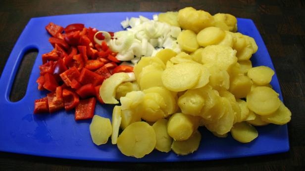 Geschnittene rote Paprika, Zwiebeln und Kartoffeln auf einem blauen Schneidebrett.