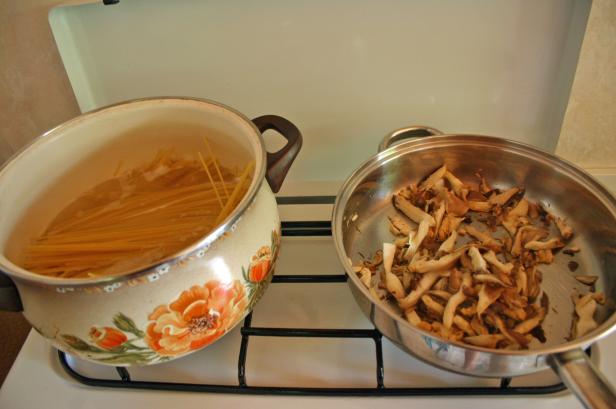Auf einem Herd stehen ein Topf mit kochender Pasta und eine Pfanne mit Pilzen.