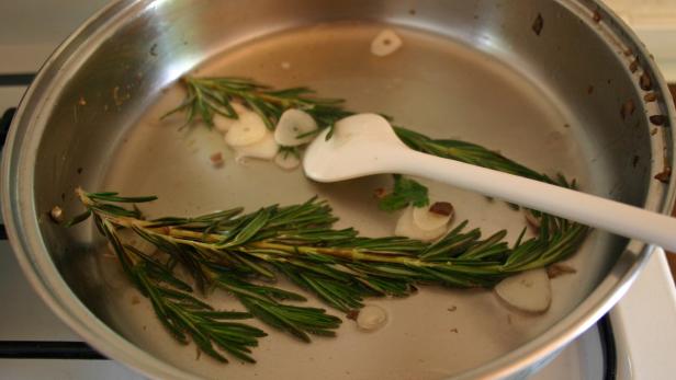 Rosmarinzweige und Knoblauchscheiben liegen in einer Edelstahlpfanne.