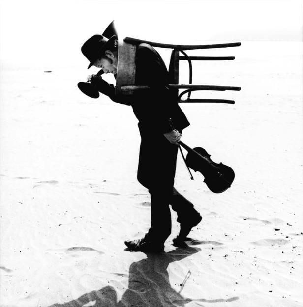 Ein Mann in Anzug und Hut geht mit einem Stuhl auf dem Rücken und einer Geige in der Hand am Strand entlang.