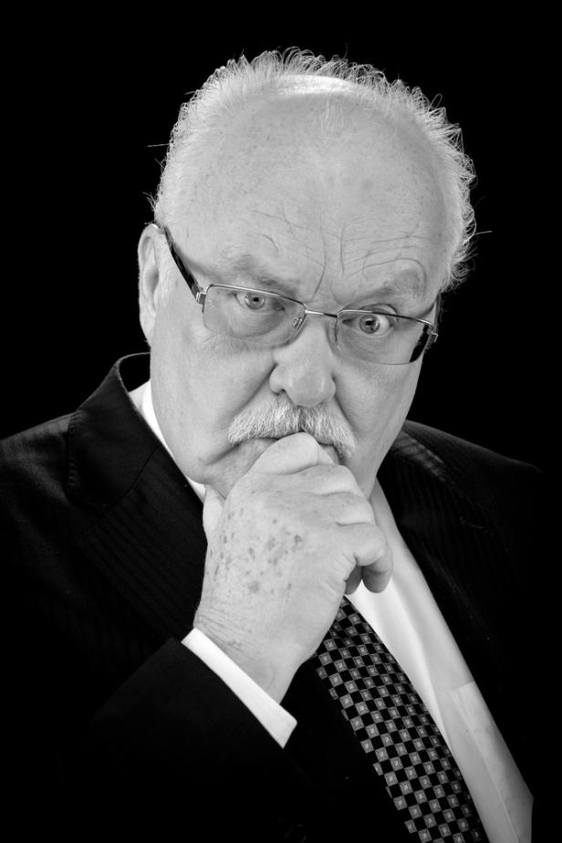 Ein Mann mit Brille und Anzug in Schwarzweißaufnahme betrachtet etwas nachdenklich.