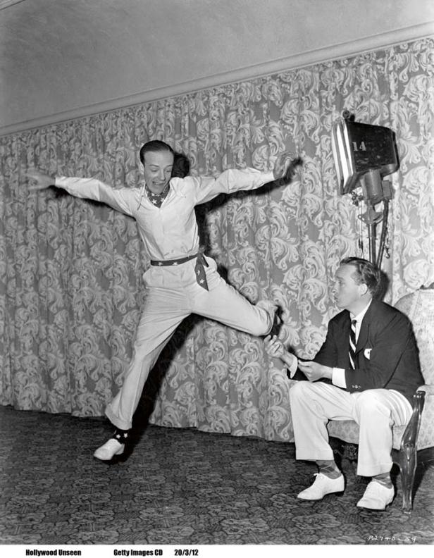 Fred Astaire springt vor einem Mann, der auf einem Stuhl sitzt.