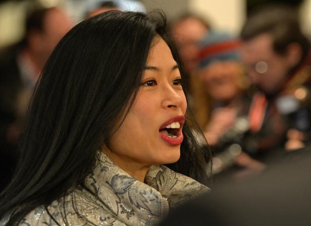 Eine Frau mit dunklen Haaren spricht vor einer unscharfen Menschenmenge.