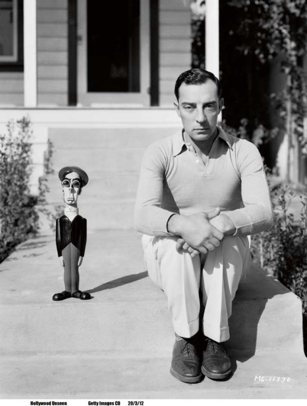 Buster Keaton sitzt neben einer hölzernen Karikatur seiner selbst auf einer Treppe.