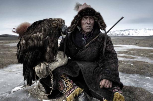 Ein mongolischer Adlerjäger sitzt mit seinem Steinadler in einer kargen Landschaft.