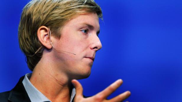 Ein Mann mit blonden Haaren und Mikroport spricht vor blauem Hintergrund.