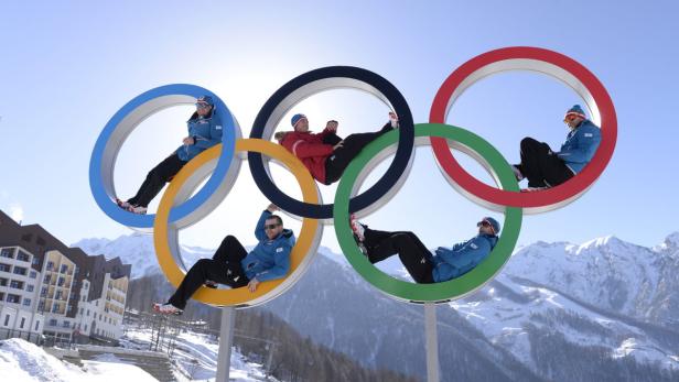 Mehrere Personen posieren in den olympischen Ringen vor einer verschneiten Bergkulisse.