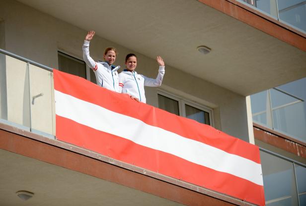 Zwei Sportlerinnen winken von einem Balkon mit einer österreichischen Flagge.