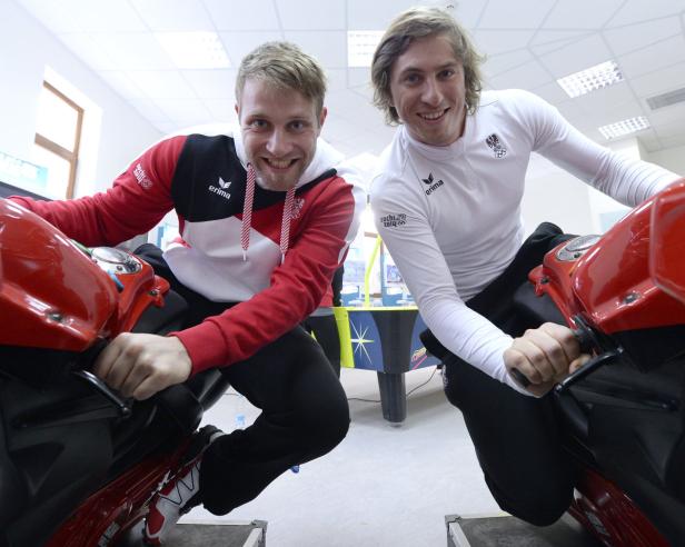Zwei Männer sitzen auf Motorradsimulatoren und lächeln in die Kamera.