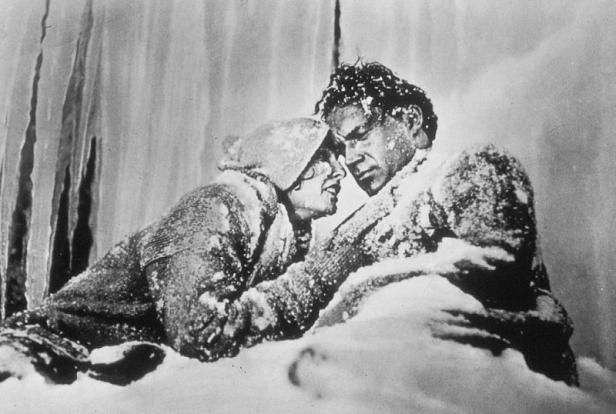 Ein Mann und eine Frau kauern verschneit in einer eisigen Umgebung.