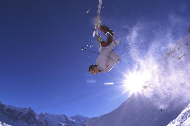 Ein Skifahrer macht einen Trick in der Luft vor blauem Himmel und schneebedeckten Bergen.