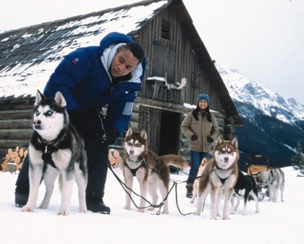 Ein Mann und eine Frau mit einem Husky-Schlitten vor einer Holzhütte im Schnee.