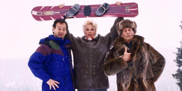 Drei Personen, darunter eine Frau mit blonden Haaren, halten ein Snowboard in die Höhe.