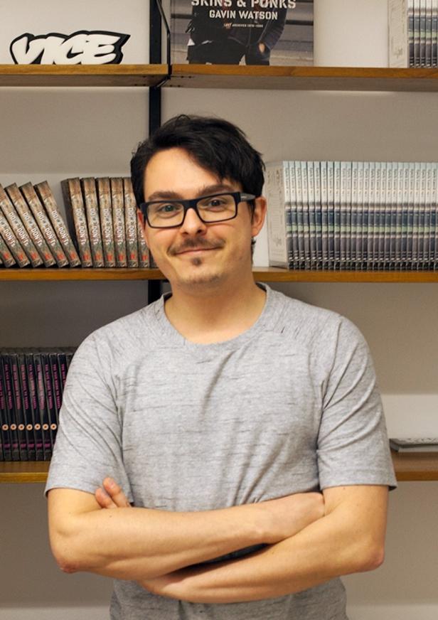 Ein Mann mit Brille und verschränkten Armen steht vor einem Bücherregal.