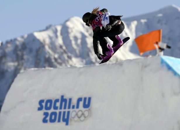 Eine Snowboarderin springt vor einer Bergkulisse über eine Rampe mit dem Aufdruck „sochi.ru 2014“.