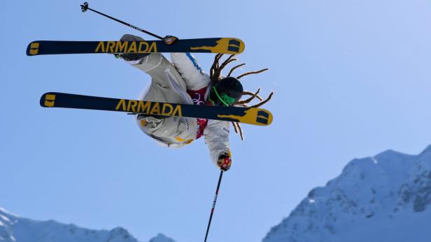 Ein Skifahrer mit Dreadlocks springt vor einem blauen Himmel und schneebedeckten Bergen.