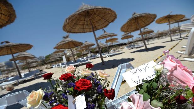 Blumen und ein Schild mit der Aufschrift „Why? Warum?“ an einem Strand mit Sonnenschirmen.