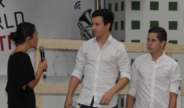 Eine Frau spricht mit einem Mikrofon zu zwei jungen Männern vor einer Wand mit Dekorationen.