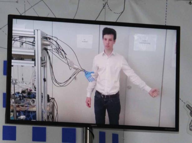 Ein junger Mann steht neben einem Roboterarm, der eine blaue Hand trägt.