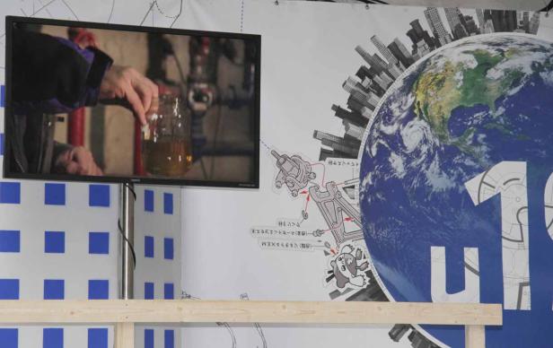 Auf einem Bildschirm ist zu sehen, wie eine Person einen Gegenstand in ein Glas mit einer Flüssigkeit taucht. Im Hintergrund ist eine Weltkugel abgebildet.