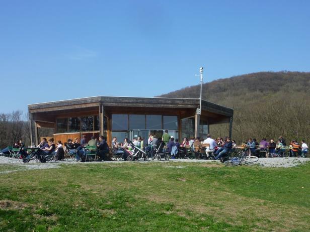 Viele Menschen sitzen an Tischen vor einem modernen Holzgebäude auf einer Wiese.