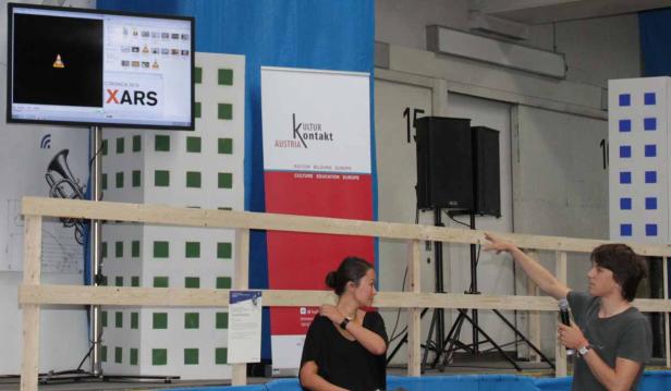 Eine Frau und ein Mann stehen vor einer Bühne mit dem Logo von „KulturKontakt Austria“.