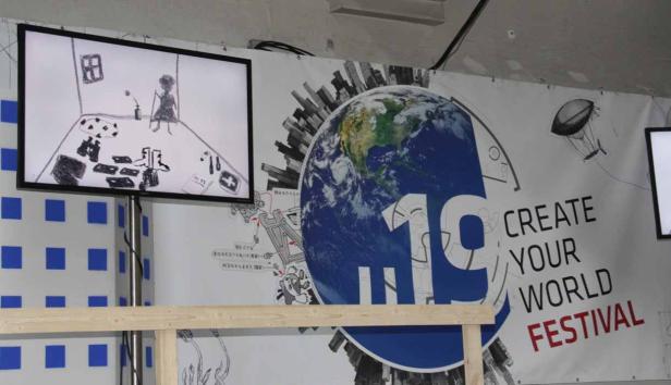 Auf einem Bildschirm ist eine Kinderzeichnung zu sehen, daneben ein Banner für das „Create Your World Festival“.