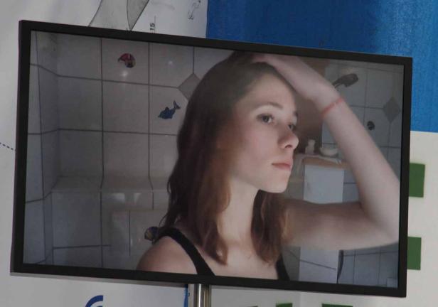 Eine junge Frau ist auf einem Bildschirm vor einer Badezimmerkulisse zu sehen.