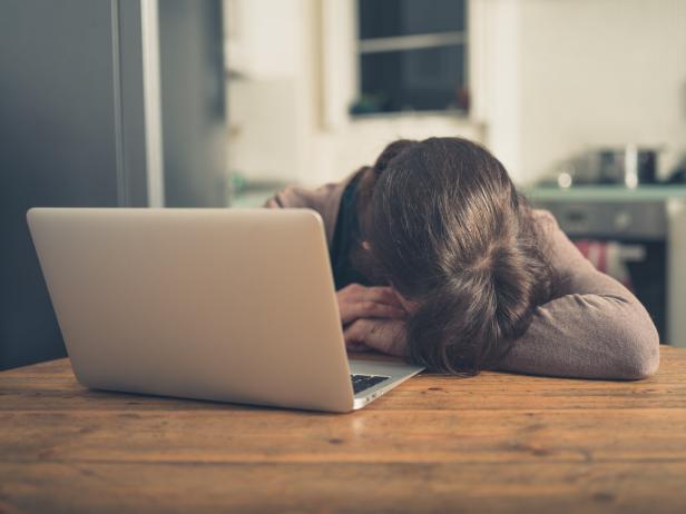 Eine erschöpfte Frau liegt mit dem Kopf auf einem Holztisch vor einem Laptop.