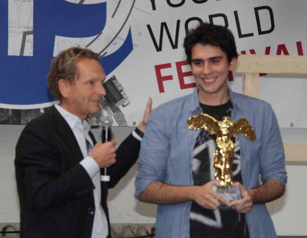 Ein junger Mann hält eine goldene Statue, während ein älterer Mann mit Mikrofon daneben steht.